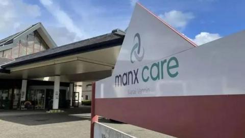 A Manx Care sign in front of the entrance of Noble's Hospital. The sign has blue, green and grey writing and the Manx Care logo. The entrance has revolving doors with a long canopy held up by pillars.