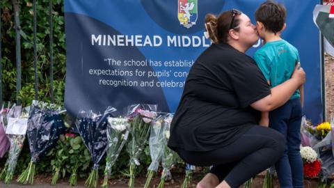 A woman bent down kissing her young son on the cheek outside Minehead Middle School. There are several bouquets of flowers left in tribute to a pupil that died.