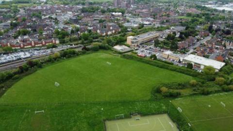 An aerial view of a town with sports pitches at the bottom and a college campus to the right. More buildings can be seen in the distance.