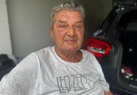 An older white man, wearing a grey t-shirt with Led Zeppelin written across the front, sits in front of a car in what appears to be a private garage