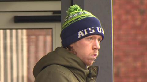 William Brown. He is wearing a green and navy bobble hat and a khaki green coat. 