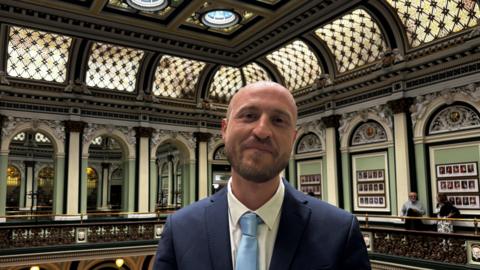 A man in a suit stands inside a council chamber