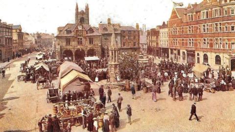 A picture of the market in Peterborough's Cathedral Square with a monument in the middle and crowds of people around covered market stalls.