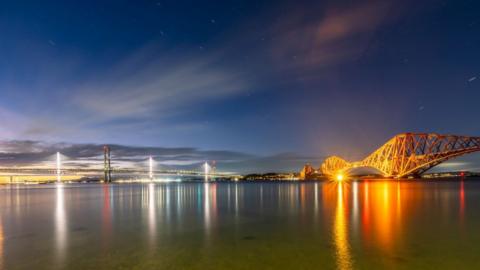 The Forth Bridge and Queensferry Crossing taken with a long exposure. The bridges are lit up and the lights are reflected in the river