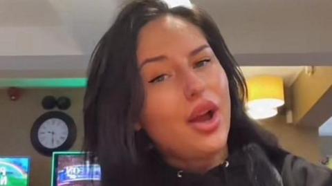 A woman with long dark hair poses in a pub, with her mouth open and a clock in the background. 