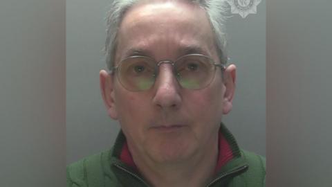 Police custody photo of Andrew Pennington, who has receding grey hair and blue eyes. He is wearing metal-framed specs on his face, a green fleece with a red trim on the collar.