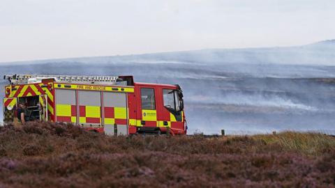 A fire engine parked on Langdale Moor. Heather can be seen in the foreground, with smoke in the distance.