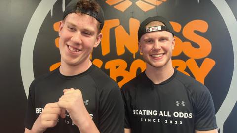 Shay and Bobby are both wearing black T-shirts and caps facing backwards. They are standing in front of a black and orange Fitness By Bobby logo.