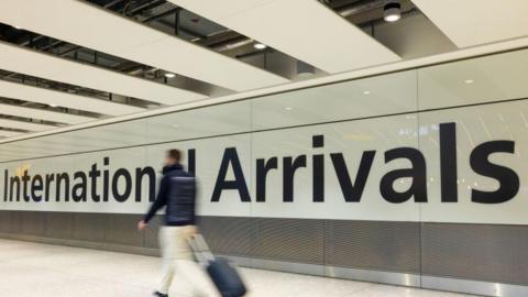 An airport corridor with the words 'International Arrivals' printed in large black letters onto a white wall. A man can be seen walking in front of the sign with a wheeled suitcase, beige trousers and a dark jacket, but he is slightly blurred to indicate the fact he is moving quickly. 