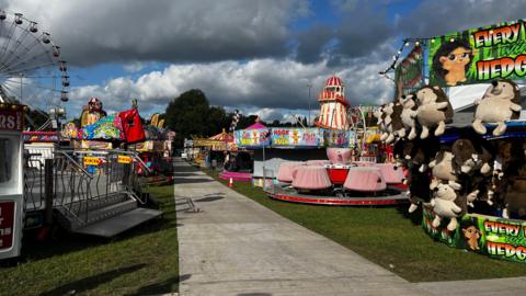 Goose Fair in 2023, with a number of fairground rides on a grassy field. At the back is a helter-skelter, and nearer is a teacups ride and a stall where people can win a hedgehog.