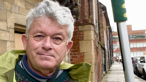 Alan Platt standing in front of an old building in Gloucester. He has grey hair and is wearing a bright yellow jacket and carrying a cleaning pole. 