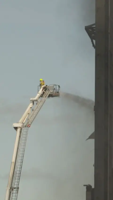 A fire fighter on a crane in a distance pouring a jet of water on to a black power station tower