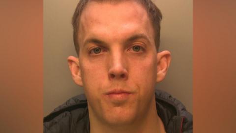 A mug shot of a man with short, brown hair. He is wearing a black coat.
