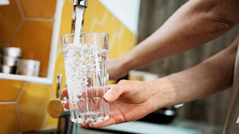 A hand filling up a glass of water at a tap in a kitchen with yellow tiles. 