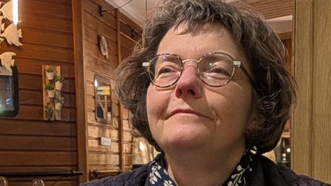 This picture shows a person seated at a table inside what looks like a cosy, warmly lit restaurant or café with a strong wooden interior. The space has wood-panelled walls, wooden ceiling slats and soft hanging lights that give the scene a relaxed, intimate feel. Decorative details are visible in the background, including framed items on the walls and small ornaments hanging from the ceiling.