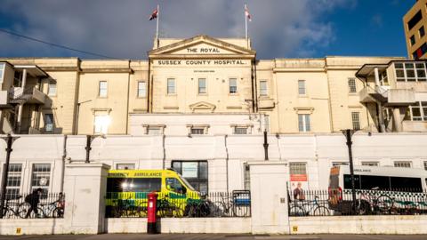 The outside of the Royal Sussex County Hospital, which is an old, white Victorian building with a Great Britain and England flag flying above the main part of the building. "Royal Sussex County Hospital" is in black text on the building.