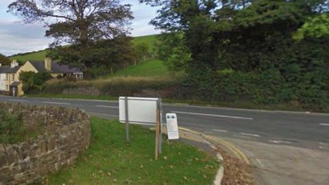 The junction at the White Hoe pumping station, showing the road running past it. There are green field and bushes across the road and a stone wall to the left next to a grass bank.