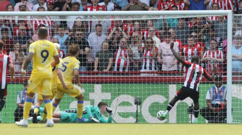 A picture from the opposite end of the pitch shows Yoane Wissa arriving unmarked at the far post to score the winner for Brentford against Crystal Palace. Goalkeeper Dean Henderson, wearing all green, is on the floor while Brentford fans behind the goal start to celebrate. 