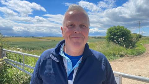 A man with grey hair, dressed in a blue zip-up jacket over a white and blue top, stands by a metal farm gate along a dirt track, with fields stretching into the distance and power lines visible under a bright, cloudy sky.