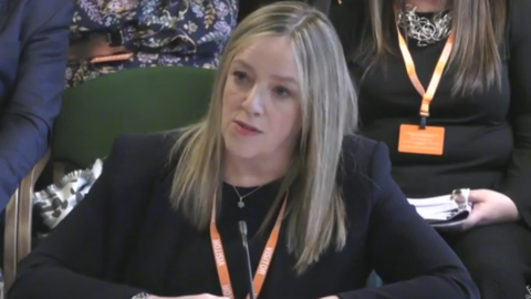 Susan Davy is sitting with her hands folded on the desk in front of her. She is wearing a navy suit and an lanyard. She has long blonde hair and looks stern. Behind her are other seated people whose faces cannot be seen.