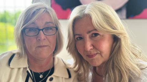 Left to right: Helen and Joanne. Helen has blonde bobbed hair and is wearing glasses, a cream and white jacket and a silver pendant around her neck. Joanne has shoulder length blonde hair and is wearing a white shirt.