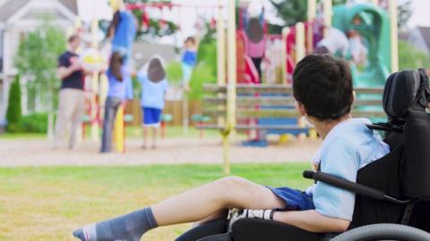 A young boy sitting in a wheelchair looking towards children playing in a playground