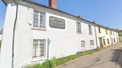 Google Street View image of The Cross Inn in Copplestone on a sunny day. The building has grey walls and has two other buildings attached to it. A sign saying "The Cross Free House - Real ALes and Fine Wines" is on the wall.