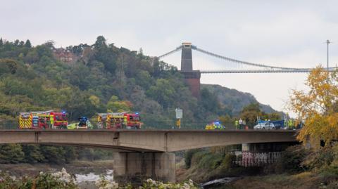 Two fire engines and two police cars are blocking the road on a concrete bridge. Their blue lights are on. A blue screen has been put up while emergency services work and a silver car can be seen on one side of the bridge. In the background are trees on the side of Avon Gorge and part of Clifton Suspension Bridge is visible in the distance.