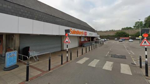 The view outside a closed Sainsbury's in Luton, from the car park. Its orange logo is above the door and the shutters are down. There is an abandoned trolley outside the front and very few cars in the car park. There is a pelican crossing in the foreground.