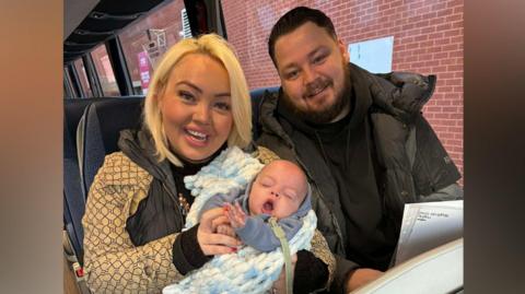 Amber and Josh Webster with their son Jude. They are sat on two seats on a bus and Amber is holding Jude. Amber has blond hair in a bob and is wearing a beige jacket. She is smiling and holding Jude, who is wearing a grey hoodie and is wrapped in a chunky knitted blue and white blanket. His eyes are shut and he is yawning. Josh has short dark hair combed back and blue eyes. He is wearing a black hoodie and black puffer jacket. He is smiling at the camera.