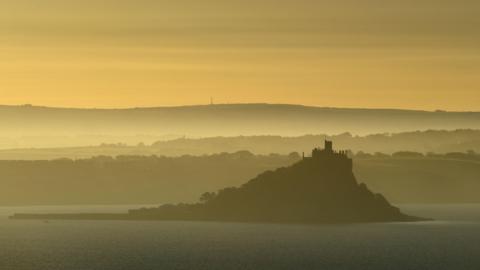 St Michael's Mount viewed across the bay during a yellow sunrise. The land behind it is swathed in mist and the castle is in silhouette.