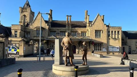 Lincoln Central Station a two-storey of light stone with pitched roofs, chimneys and gothic tower. In the foreground is a statue of Victorian mathematician George Boole, depicting Boole lecturing two children
