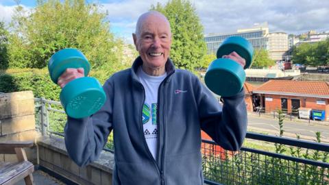 92-year-old Ken Head smiles as he lifts two hand weights, wearing a blue fleece top on his balcony which looks out across Bristol.