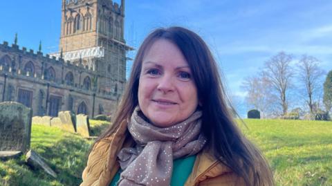 A brunette woman is smiling in front of a church with scaffolding on its spire. She wears a green top with a flower, a yellow puffer jacket, and a brown sparkly scarf. The sun is shining and the sky is clear blue.