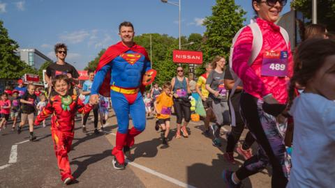 Children holding hands with adults start the Great Bristol run. Some are wearing sportswear whilst others are dressed as superheroes