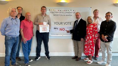 Sir Geoffrey Clifton-Brown pointing to a whiteboard with a sign attached to it that says "Further development of Moreton-in-Marsh" "Your vote - 96 per cent NO). He is joined by seven other people, including councillors.