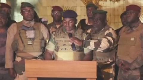 Army officers stand in front of podium while one man at front speaks into microphone