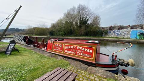 A red narrowboat with writing on it reading "Dane" and "Mersey Weaver and S.C.C.Co Ltd General Carriers" in a canal. A picnic table, a black sign and grass are on one side of the canal while a wall and trees are on the other side.