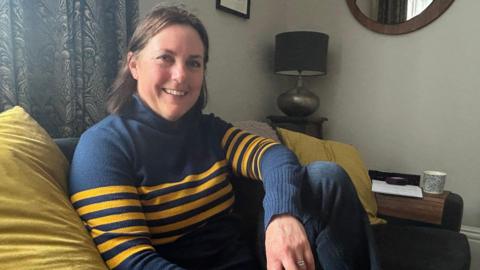 A woman with dark brown shoulder-length hair sits in a living room on a sofa with green-yellow cushions on it. She is wearing blue jeans and a dark blue sweater with horizontal yellow striped across the body and arms. She is smiling at the camera.