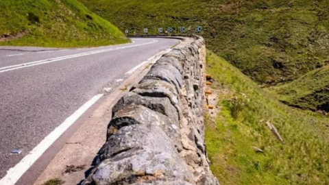 A stretch of the A57 Snake Pass showing a sudden drop at the side of a rocky wall. The wall is almost the same level as the road and there is a sharp bend towards the end of the road.