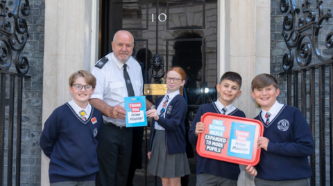 Four 11-year-old schoolchildren, three boys and a girl in blue uniforms, hand a card to a police officer on the steps of Number 10