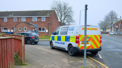 A blue and yellow marked police crime scene investigation van parked on the side of a road beside a row of terraced red brick houses. A black car is parked in front of it. The sky is grey.
