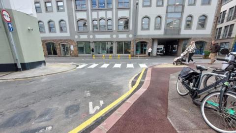 A mock-up of a zebra crossing at the end of a town centre junction. It is surrounded by quite high buildings, and there are pedestrians and bicycles nearby. 