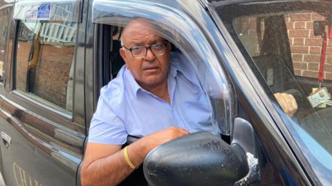 An Asian man wearing glasses and with bald head and grey hair on the sides, is wearing a pale blue short leave shirt. He is leaning his right elbow out of the drivers side window of a black cab window.