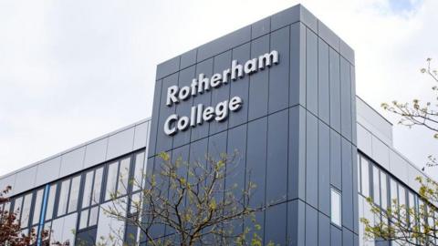 Against a cloudy sky, there's a tall grey block building. Windows run along two sides. At the corner between the two sides, a taller dark grey block has the words Rotherham College on it, in large white letters. There are trees in leaf at the far left and right of the picture and in the middle, which reaches up to just below the name of the college
