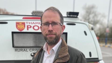 Matthew Barber is standing in a green, waxed jacket in front of a white police van.