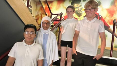 Four children, two boys and two girls, standing in a stairwell. They are wearing summery school uniform. Behind them is a large mural depicting a photo of a footballer.