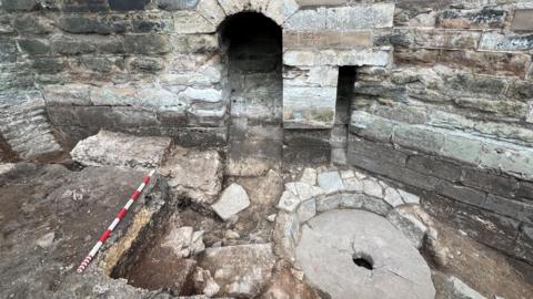 A stone circle with grey blocks around it. The circle has a small hole in the middle and there is a red and white striped measuring stick also laid across the stonework.