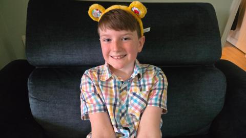 Taylor wearing a gingham, multi-coloured shirt and Pudsey ears smiling at the camera while sitting in a large dark armchair.