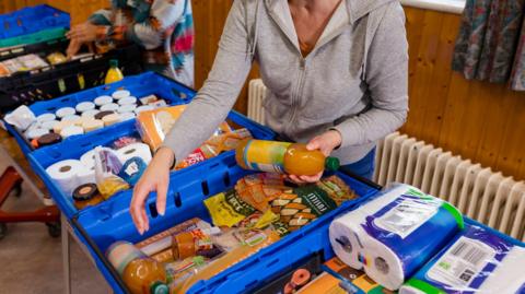 A medium shot of a food bank being run by volunteers at a community centre. Food that has been donated is lined up in crates on the table.